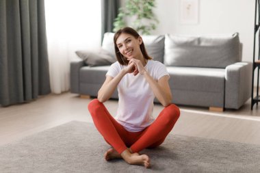 Beyaz tişörtlü, kırmızı taytlı, çapraz bacaklı, pozitif genç bir kadının ön görüntüsü. Çekici beyaz kadın, rahat bir dairede sabah egzersizleri arasında dinleniyor..