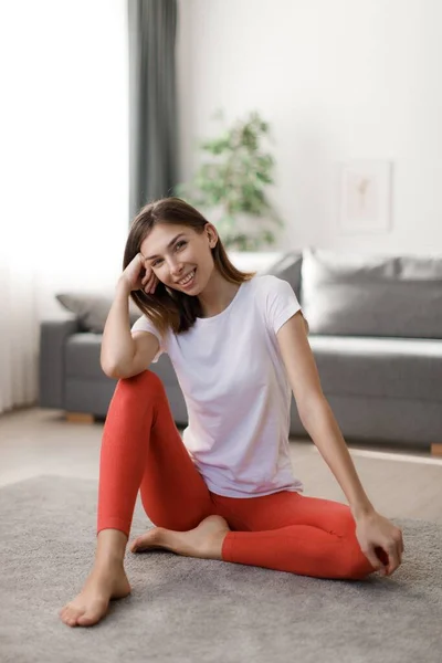 stock image Stunning young woman in sport clothes sitting on floor and smiling sincerely. Caucasian dark haired lady feeling satisfaction after morning workout at home.