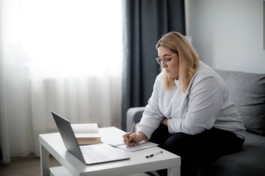Beyaz kapüşonlu beyaz bir öğrenci evde dinlemek için taşınabilir laptop kullanıyor. Gözlüklü güzel kadının yan görüntüsü Online dersten bazı cümleleri not alıyor.