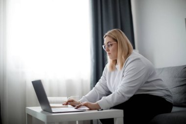 Gözlüklü, kendine güveni tam bir kadın oturma odasındaki kanepede otururken modern dizüstü bilgisayarda ders izliyor. Araştırmalar için farklı bilgi kaynakları kullanan şişman genç bir kadın..