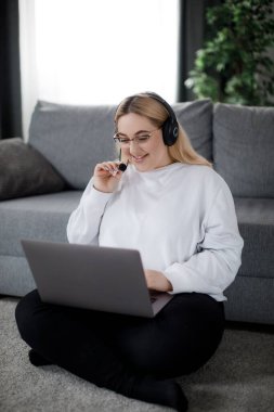 Ayrıca kulaklıklı kadın kablosuz bilgisayarla yerde oturuyor ve video görüşmesi yapıyor. Mutlu genç sarışın evde otururken online eğitimin tadını çıkarıyor..