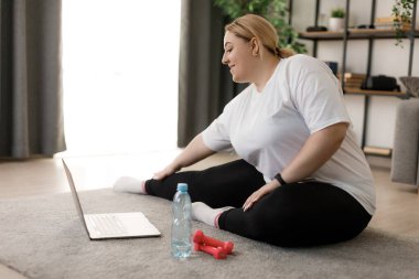 Pozitif kıvrımlı kadın, yerde oturup modern dizüstü bilgisayardan online ders izlerken esneme egzersizleri yapıyor. Yurt içi çalışma teknolojisi. Kilo verme konsepti.