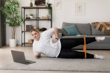 Ayrıca beden ölçülerinde bir kadın antrenman sırasında lastik bant kullanıyor. Spor elbiseli bir bayan yerde uzanıyor ve taşınabilir dizüstü bilgisayardan spor videosu izliyor..