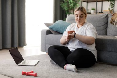 Ayrıca spor ayakkabılı, bileğinde spor takip cihazı olan yumuşak halıda oturan ve modern dizüstü bilgisayar kullanan bir kadın. İç egzersizler sırasında kalp atışlarını kontrol eden beyaz sarışın..