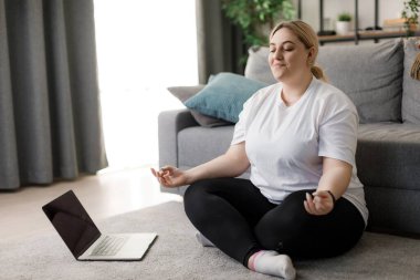 Fazladan ağırlığı olan huzurlu genç bir kadın lotus pozisyonunda oturuyor ve gözleri kapalı meditasyon yapıyor. Kafkas kadın sarışın evde yoga pratiği için modern laptop kullanıyor..