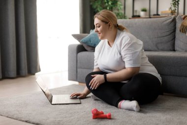 Oturma odasında halıya oturmuş spor videosu izlemek için modern dizüstü bilgisayar kullanan aktif kıyafetli bir kadın. Yurtiçi eğitim ve teknoloji kavramı.