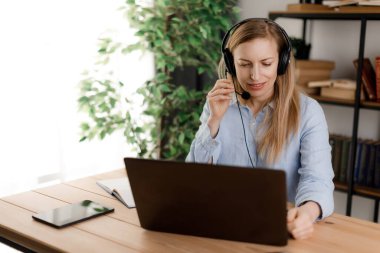 Kulaklıklı, mikrofonlu konsantre bir kadın evde çevrimiçi toplantı yaparken laptop ekranına bakıyor. Çevrimiçi iş ve modern teknoloji kavramı.