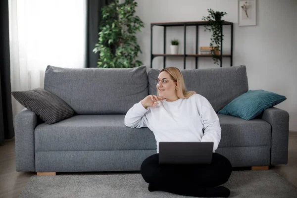 Düşünceli öğrenci oturma odasında modern kanepenin yanında otururken gülümsüyor ve yan bakıyor. Çevrimiçi kursları izlemek için dizüstü bilgisayar kullanan gözlüklü mutlu şişman kadın..