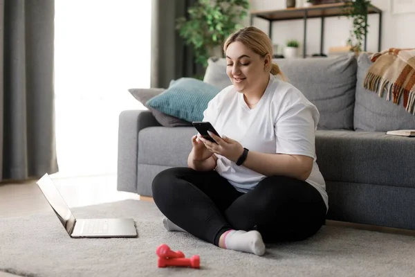 Keyifli, geniş beden giyimli, dizüstü bilgisayarı ve dambılı olan ve modern akıllı telefondan mesaj atan bir kadın. Aletlerle ve spor aktiviteleriyle boş zaman.