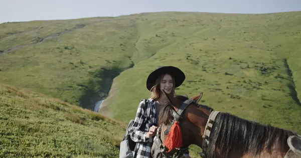 stock image In the vast greenery of the Carpathian Mountains, a female tourist, dressed in casual hiking gear and a hat, is captured adjusting the bridle of her horse. The scene showcases her readiness to embark