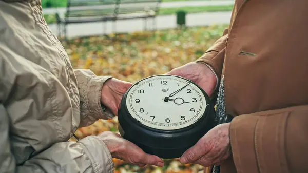 stock image Elderly man and woman looking at huge clock and walking away together in blurred autumn park