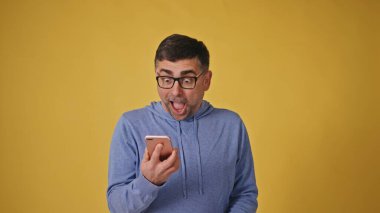 Overjoyed mature man in glasses reacting to smartphone news on yellow background. High quality 4k footage clipart