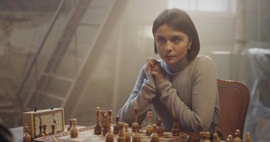 A woman in a gray sweater intensely focuses on the chessboard, strategizing her next move in a warm, vintage-inspired setting. The wooden chess pieces, classic clock, and soft ambient lighting create clipart