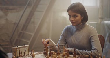 A thoughtful woman in a gray sweater deeply concentrates on her next chess move in a vintage-inspired room. The warm lighting enhances the scene, highlighting the chessboard, clock, and rustic clipart