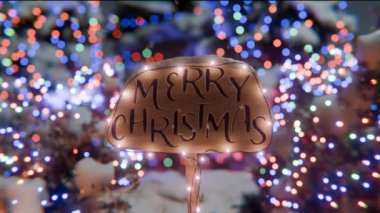Bayram ışıklarıyla aydınlatılmış mutlu noel ahşap tabelaları şenlik ışıklarıyla süslenmiş Noel ağaçlarının zemininde. Yumuşak kamera hareketi.