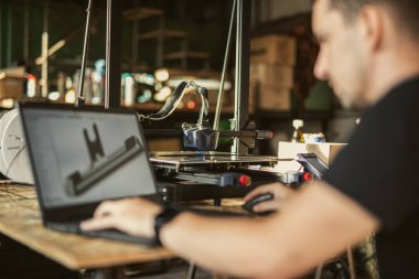 Bir mühendis, güneşli bir atölyede 3D yazıcıya basılan bir dizüstü bilgisayarda 3D bir element tasarlıyor. Resim yaratıcı süreci yakalar