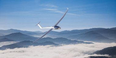 Puslu dağların üzerinden süzülen parlak bir planör berrak mavi gökyüzünün altında, dinginliğin ve özgürlüğün özünü yakalıyor..