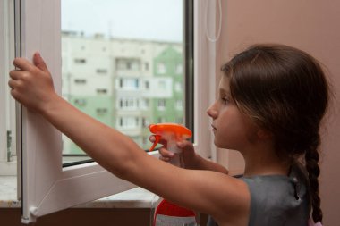 Küçük kız evde pencereleri temizliyor. Plastik sprey su şişesinden pencereye püskürtmek için..