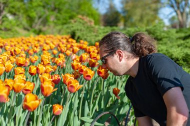 Genç adam turuncu laleleri kokluyor. Siyah güneş gözlüğü ve siyah kıyafetler..