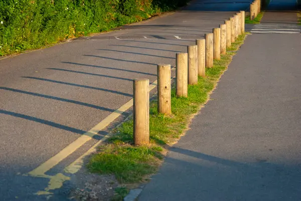 Kısa tahta direklerle çevrili asfalt bir yol. Direkler, güneş ışığının alçak açısı nedeniyle yol boyunca uzun ve keskin gölgeler oluşturuyor. Bu da ya sabahın erken saatlerinde ya da öğleden sonra olduğunu gösteriyor..