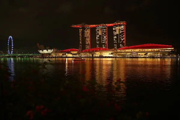 Marina Bay Sands 'e bakan Merlion Park' tan Singapur manzarası. Marina Körfezi 'nin ışıkları yüzeyinde çarpıcı yansımalar yaratırken tekneler yavaşça suda süzülüyor..