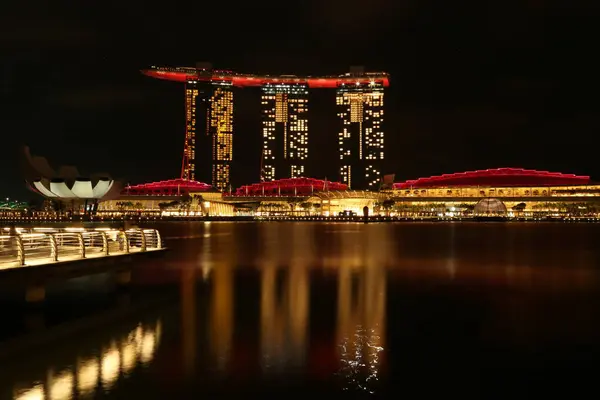 Marina Bay Sands 'e bakan Merlion Park' tan Singapur 'un gece görüşü. Marina Körfezi 'nin ışıkları yüzeyinde çarpıcı yansımalar yaratırken tekneler yavaşça suda süzülüyor..