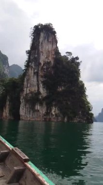 Sahne kireçtaşı kayalıkları, sakin tropikal bir göldeki zümrüt yeşili sulardan dramatik bir şekilde yükselir. Arka planda puslu dağlar gizemli bir cazibe katar, sakin ve resimsel bir doğa yaratır..