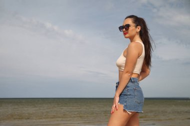 Kot şortlu kadın denizin kenarında duruyor.
