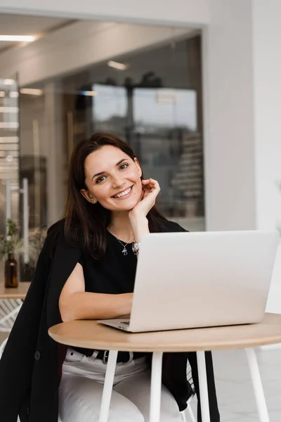 stock image Cheerful young woman programmer works remotely on laptop and try to meet deadline. Candid girl with laptop sits in white cafe, smiles and rejoices at successful work in IT company