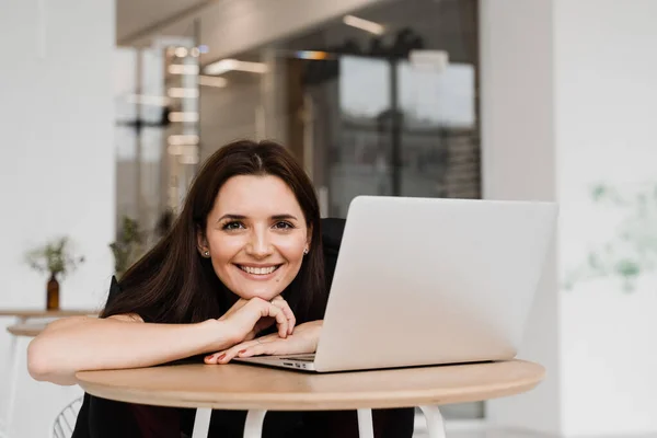 Candid girl with laptop sits in white cafe, smiles and rejoices at successful work in IT company. Cheerful young woman programmer works remotely on laptop and try to meet deadline