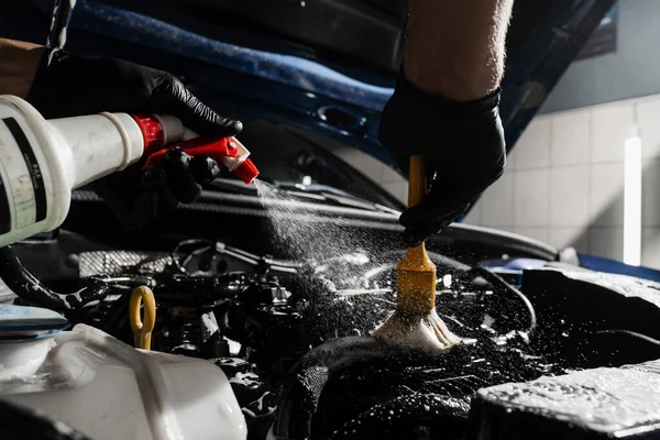 Washing car engine with spray, brush and detergent in detailing auto service. Detailing cleaning motor from dust and dirt. Spraying detergent on car engine
