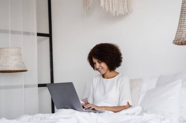 Neşeli Afrikalı kız sabahları yatak odasındaki rahat yatakta dizüstü bilgisayarla çalışıyor. Tembel Afrikalı Amerikalı kadın programcı evden kodlama yapıyor ve iş arkadaşlarıyla yatakta buluşuyor.