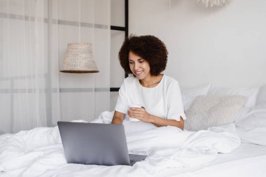 Bilgisayarlı, bir fincan kahve içen, online film izleyen ve yatak odasında internette sörf yapan mutlu Afrikalı kız. Afro-Amerikan bir kadının dizüstü bilgisayarıyla sıcak bir sabah.