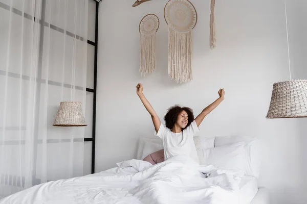 穿着睡衣的非洲女孩躺在舒适的床上 双手高举在家里 非洲裔美国女人闭着眼睛醒来 享受着早晨的愉快 — 图库照片
