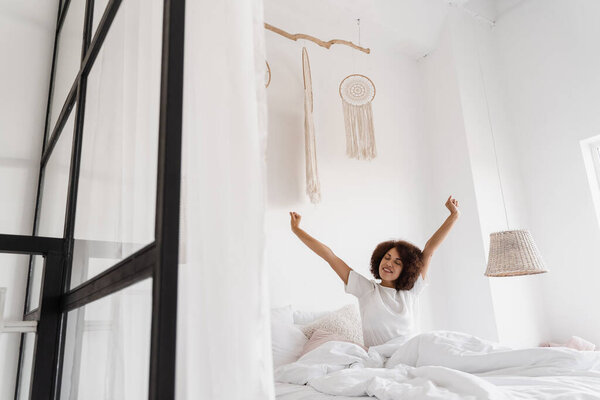 Африканская американка просыпается и наслаждается добрым утром. Африканская девушка в пижаме, сидящая в уютной постели и поднимающая руки дома
