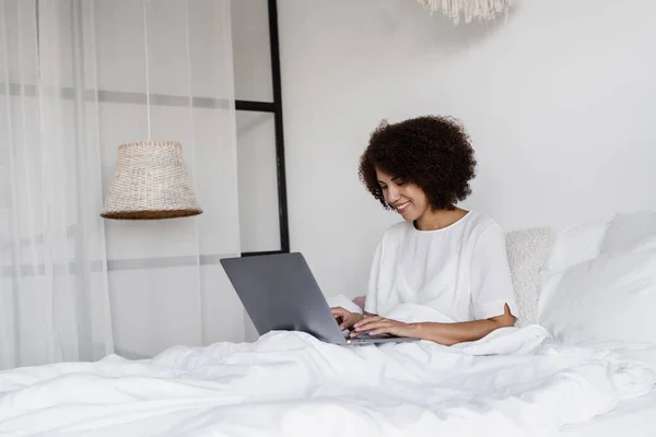 Cheerful african girl working on laptop in cozy bed in bedroom in the morning. Lazy african american woman programmer coding from home and have business meeting with colleagues in bed