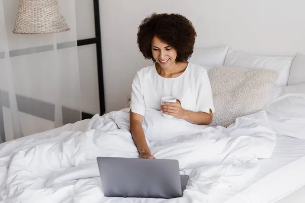Happy african girl with laptop drinking cup of coffee, watching online movie and surfing internet in bedroom at home. Cozy morning with laptop of african american woman