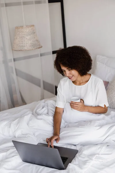 stock image Happy african girl with laptop drinking cup of coffee, watching online movie and surfing internet in bedroom at home. Cozy morning with laptop of african american woman