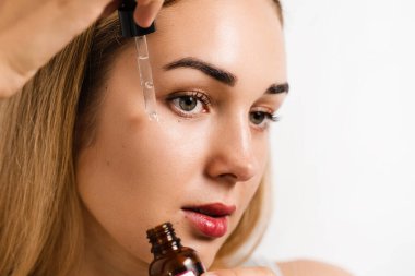 Beyaz arka planda çekici bir kız olan yüz serumu pipeti. Kız yüzüne beyaz arka planda pipetle yüz serumu uyguluyor.