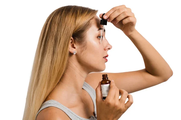 Girl applies face serum to her face with pipette on white background. Young woman makes rejuvenating cosmetic procedure for skin and face