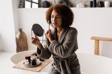 Afro-amerikan neşeli kadın güzellik spa ameliyatları yapıyor ve cilt bakımı yapıyor. Bornozlu Afrikalı kız beyaz banyoda aynaya bakıyor.