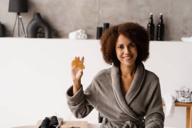 Banyoda altın hidrojel göz bantlı, peptitli ve hyaluronik asitli bornoz giyen Afrikalı bir kız. Kırışıklıkları düzeltmek için altın göz bandı tutan kadın, şişliği ya da göz altı torbalarını çıkarın.