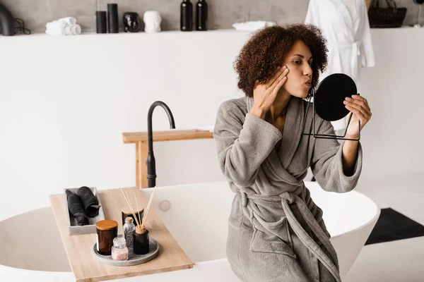 Bornozlu Afrikalı Amerikalı bir kızın güzel portresi. Çekici kadın sivilcelerine ve sivilcelerine bakıyor. Güzellik sabahı banyoda rutin bir cilt bakımı