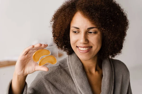 African woman holding gold eye patches for smoothing wrinkles, remove swelling or bags under eyes. Girl in bathrobe with golden hydrogel eye patches with peptides and hyaluronic acid in bathroom