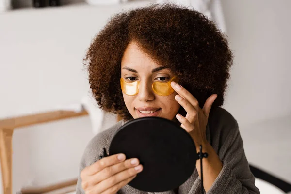 African girl in bathrobe applies gold patches to her eyes in the bathroom. Morning beauty routine. African american woman with golden patches on her eyes looks in the mirror at her skin