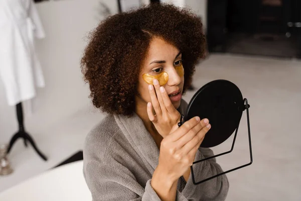 Morning beauty routine. African american woman with golden patches on her eyes looks in the mirror at her skin. African girl in bathrobe applies gold patches to her eyes in the bathroom