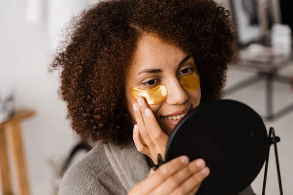 Morning beauty routine. African american woman with golden patches on her eyes looks in the mirror at her skin. African girl in bathrobe applies gold patches to her eyes in the bathroom