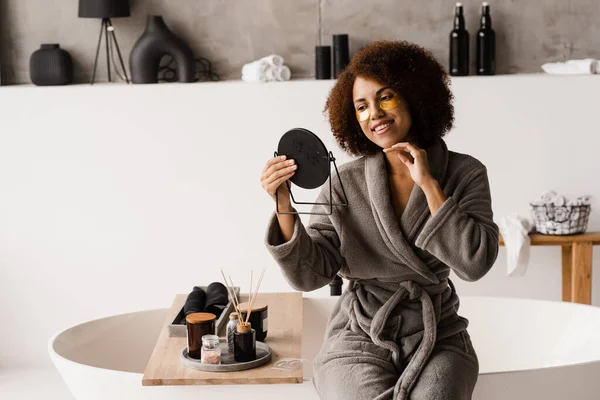 African american woman with golden patches on her eyes looks in the mirror at her skin. Morning beauty routine. African girl in bathrobe applies gold patches to her eyes in the bathroom