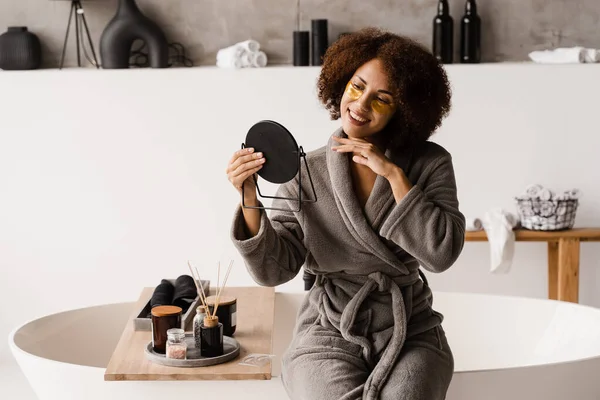 African american woman with golden patches on eyes looks in the mirror. Morning beauty routine. African girl in bathrobe applies gold patches to her eyes in the bathroom