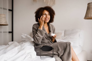 Afrikalı bir kız, cildini yatak odasındaki kuruluktan korumak için yüz nemlendirici kremi sürüyor. Yüzü nemlendirici kremle sabahları rutin güzellik yapan Afrikalı Amerikalı kadın.